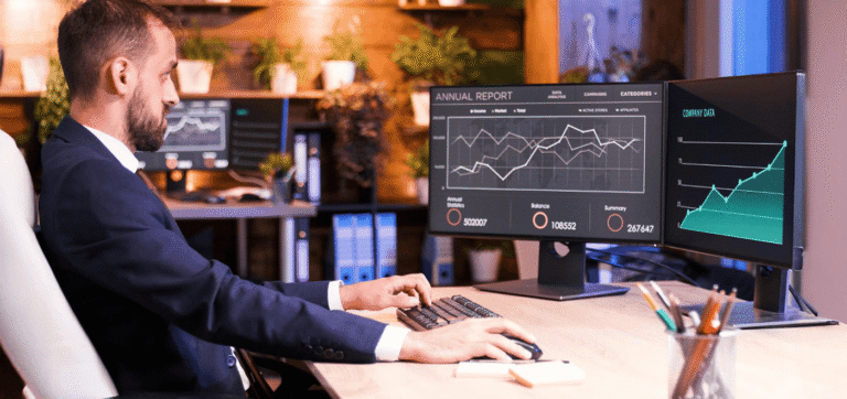 Man in a suit sits at a desk and types on a keyboard while two monitors display financial charts and an annual report; data visuals dominate the screen space.