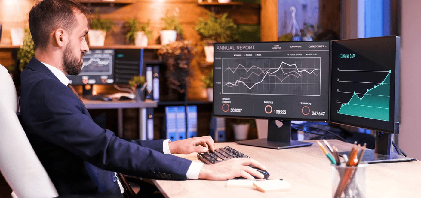 Man in a suit sits at a desk and types on a keyboard while two monitors display financial charts and an annual report; data visuals dominate the screen space.
