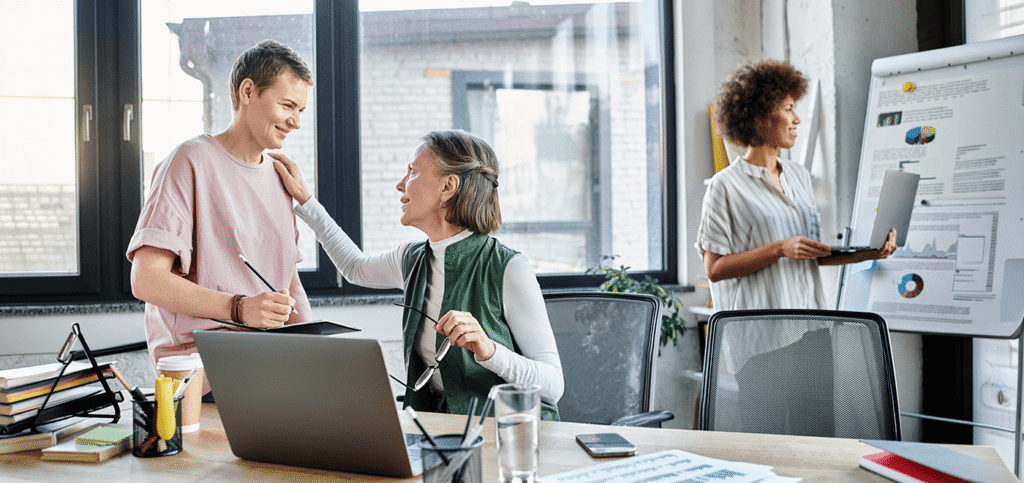 Two colleagues smile and chat at a desk while a third person presents data on a whiteboard in the background.
