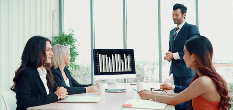 Business meeting in a bright office; presenter in a suit shows a bar chart on a monitor to three colleagues around a conference table.